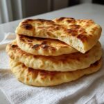 A stack of four golden-brown, fluffy pita bread rounds, with the top one broken to show the soft interior texture.