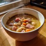 A steaming white bowl filled with hearty bean soup containing white beans, chunks of ham, and carrots, sitting on a wooden surface.