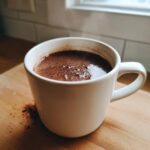 A close-up of a white mug filled with rich, dark hot cocoa mix, steaming slightly on a wooden surface.