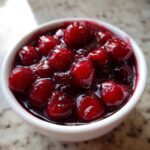 Close-up of glossy, thick cherry pie filling with whole, bright red cherries in a small white bowl.
