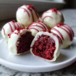 Close-up of several red velvet cake balls coated in white chocolate, with two cut open revealing the deep red interior.